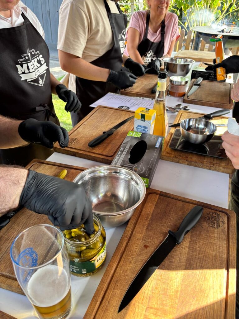 Teilnehmer mit Grillschürzen von Meck'sBBQ schneiden Zutaten an einem langen Vorbereitungstisch im Freien.