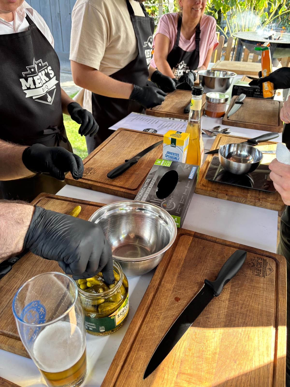 Teilnehmer mit Grillschürzen von Meck'sBBQ schneiden Zutaten an einem langen Vorbereitungstisch im Freien.